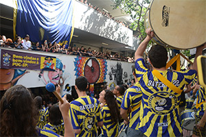 Foto de vários estudantes assistindo apresentação de grupo de alunos participantes de bateria da PUC-SP, Campus Monte Alegre. Em primeiro plano, estes alunos estão de costas